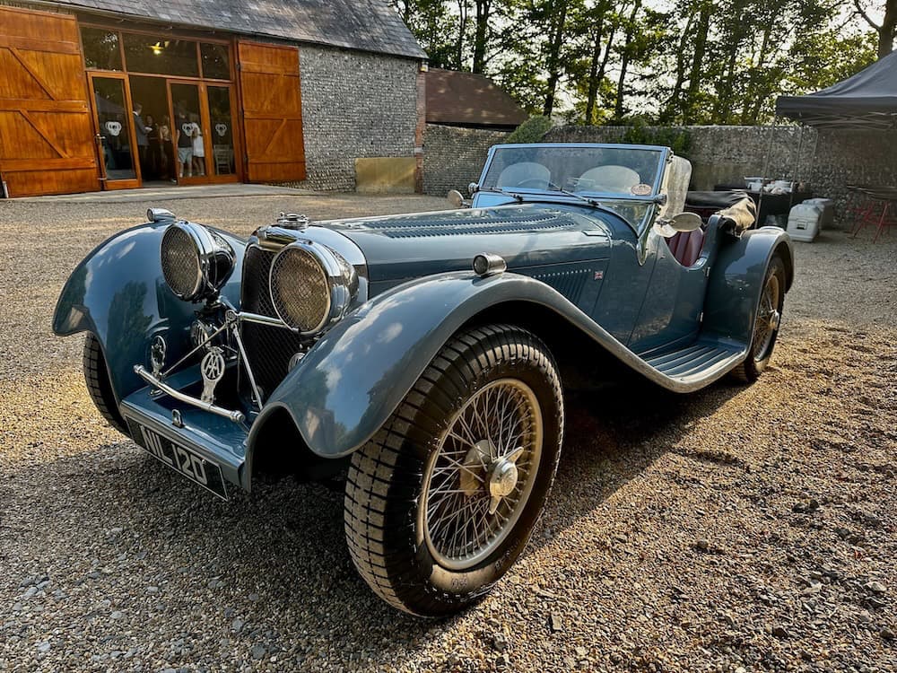 An SS100 classic car in pale blue on gravel driveway with Cissbury barn in the background